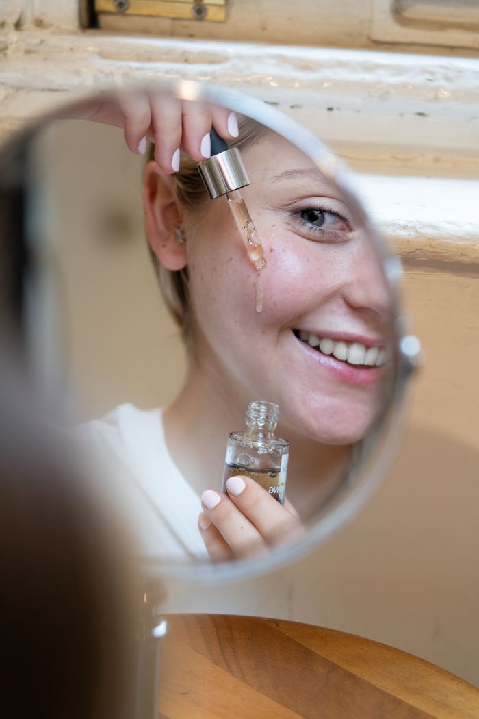 Smiling woman applying face serum with dropper seen in mirror reflection.